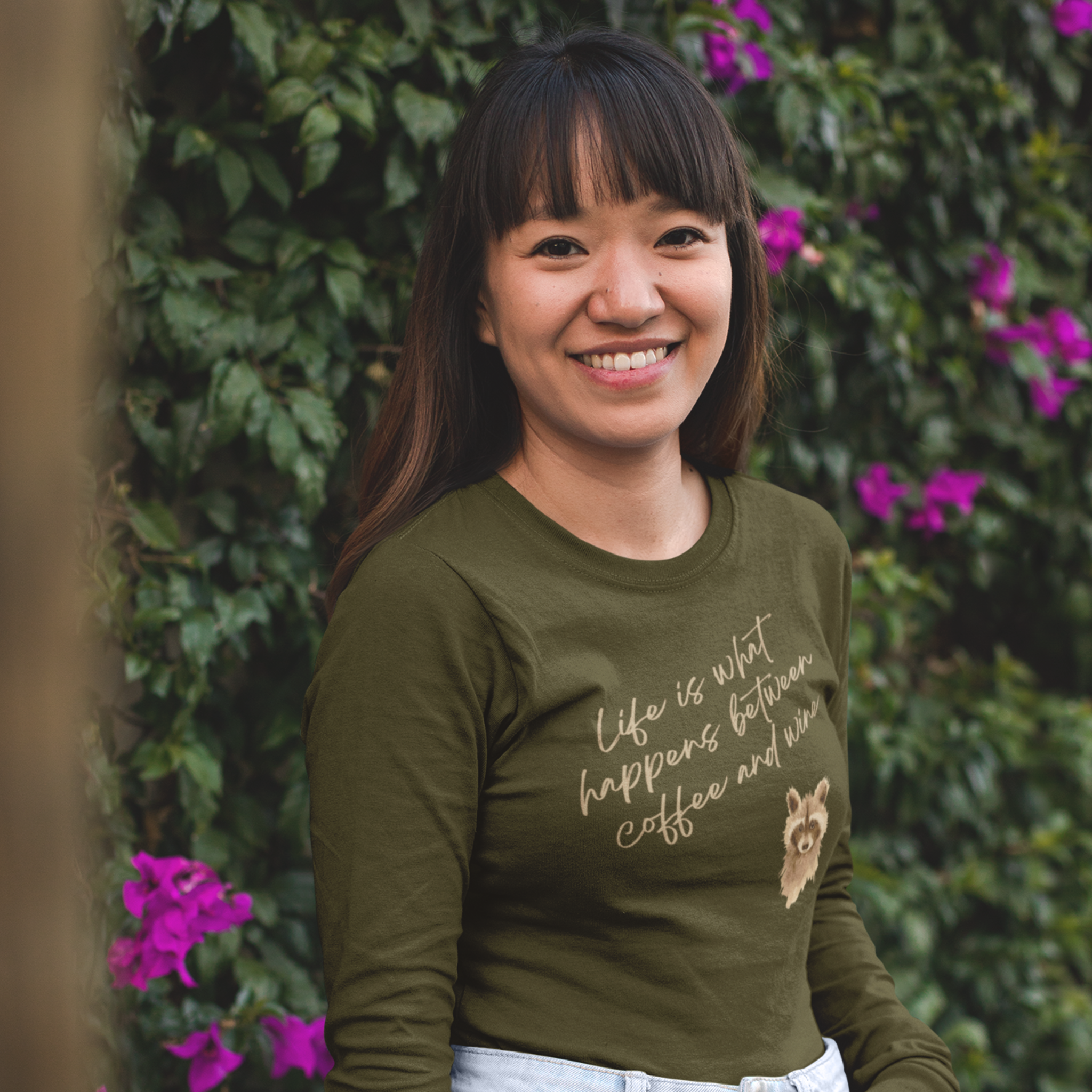 Woman wearing a green shirt with text and a bear graphic, standing in front of greenery with purple flowers.